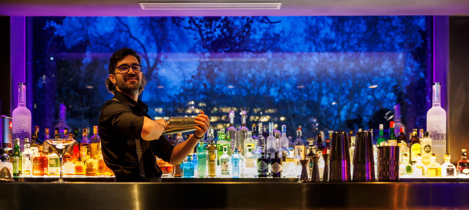 A male bartender making a cocktail in front of a glowing blue background bar shelf of spirits.