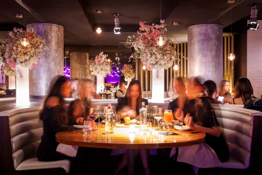 Group of diners at a glowing round table in a modern restaurant, with blurred motion, hanging floral arrangements, and ambient purple lighting in the background.