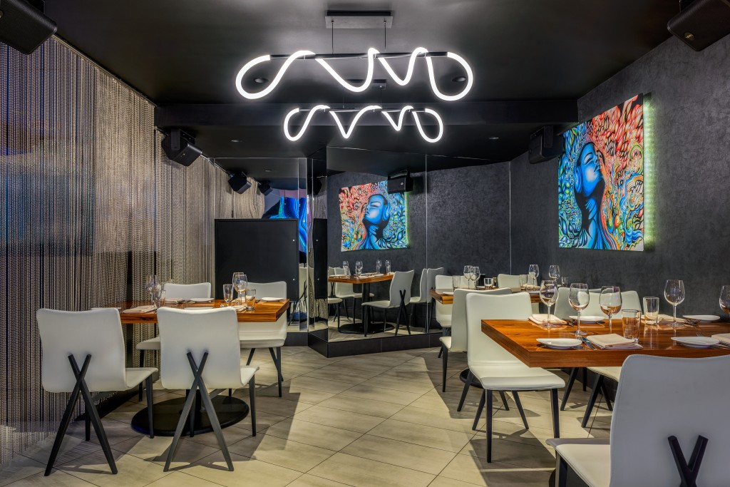 STK Washington interior featuring dark walls and ceiling, highlighted by a sculptural light fixture with looping, neon like white tubing. Tables are set with glassware and napkins, surrounded by white chairs with black legs.